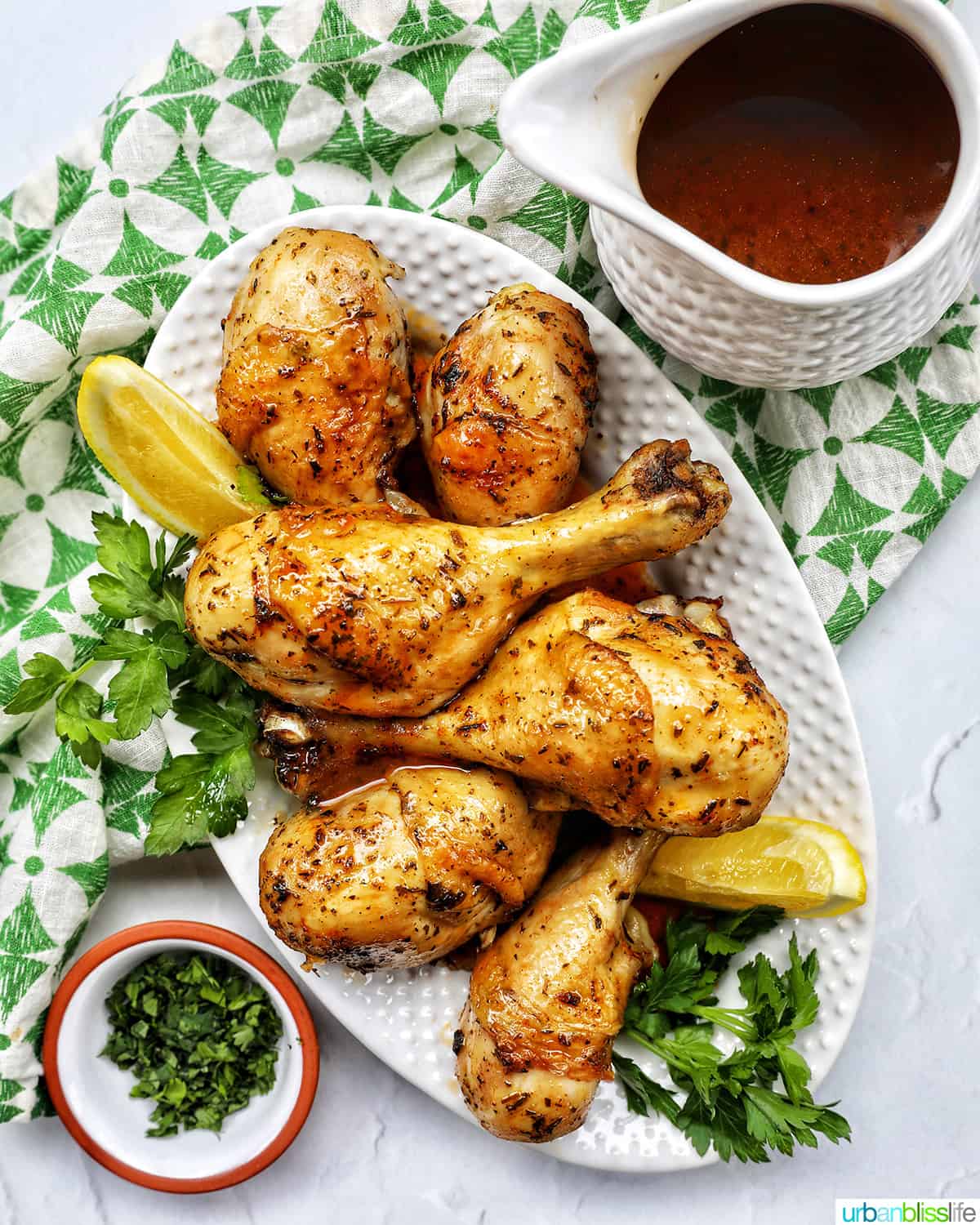 Stack of chicken drumsticks with seasoning and garnished with parsley and side of lemon wedges on bed of herbs and side of gravy.