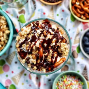 ice cream parfait topped with granola, chocolate syrup, mini chocolate chips on a colorful kitchen towel surrounded by sides of more toppings.