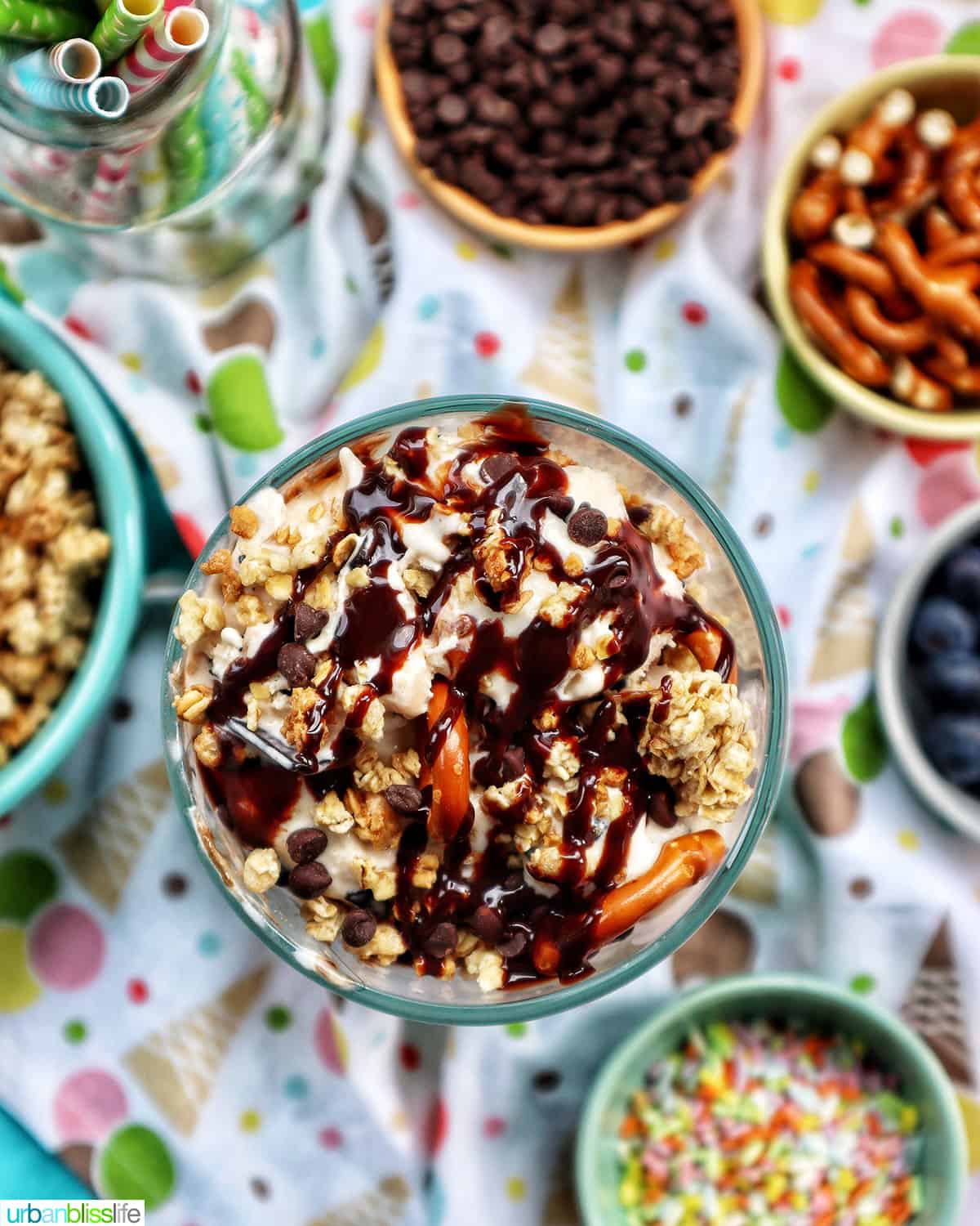 ice cream parfait topped with granola, chocolate syrup, mini chocolate chips on a colorful kitchen towel surrounded by sides of more toppings.