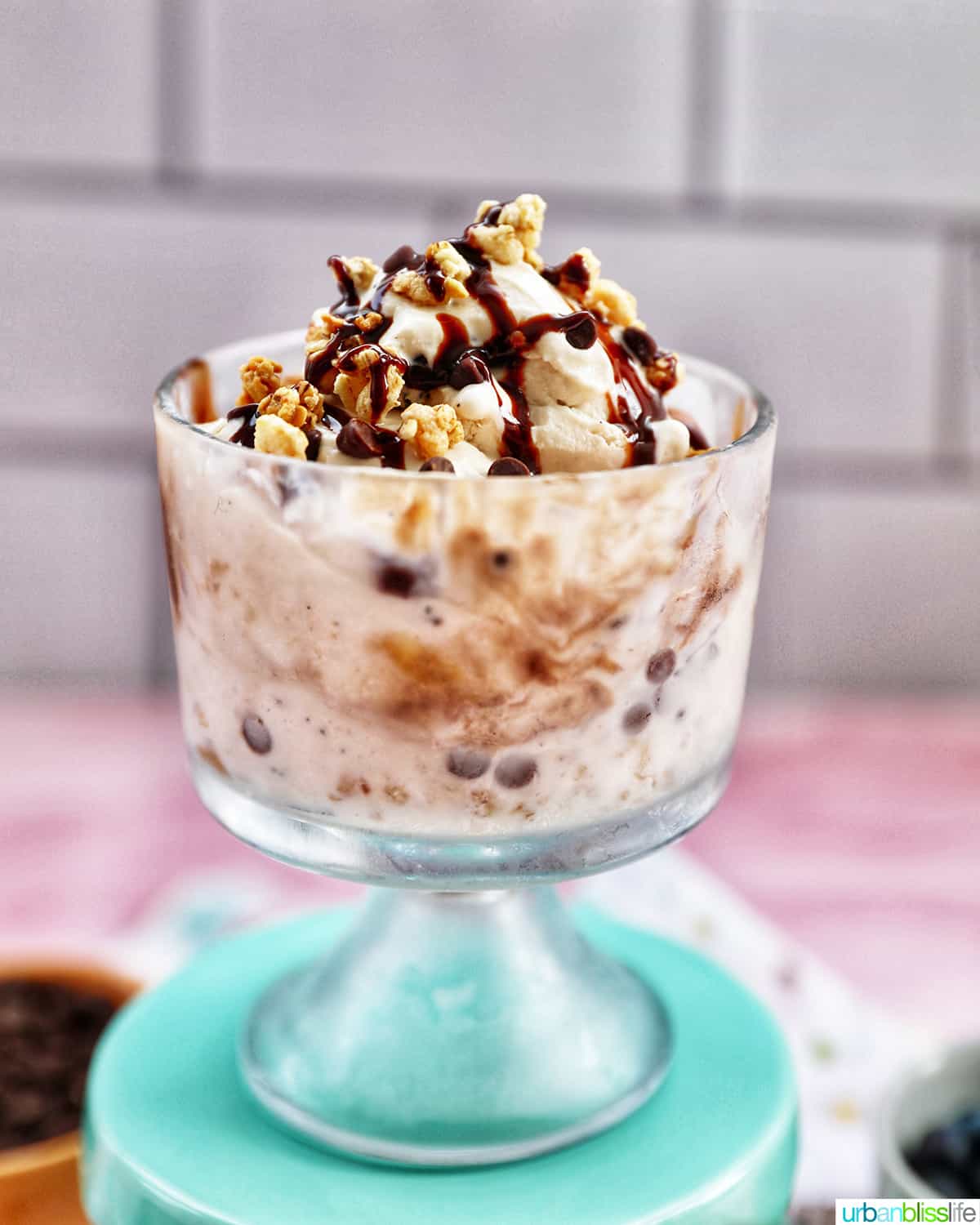 parfait bowl with ice cream parfait topped with granola, chocolate syrup, mini chocolate chips on a colorful kitchen towel.