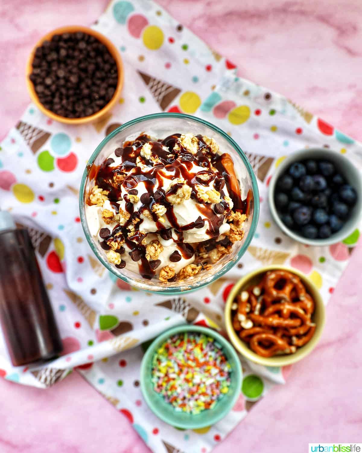 ice cream parfait topped with granola, chocolate syrup, mini chocolate chips on a colorful kitchen towel surrounded by sides of more toppings.