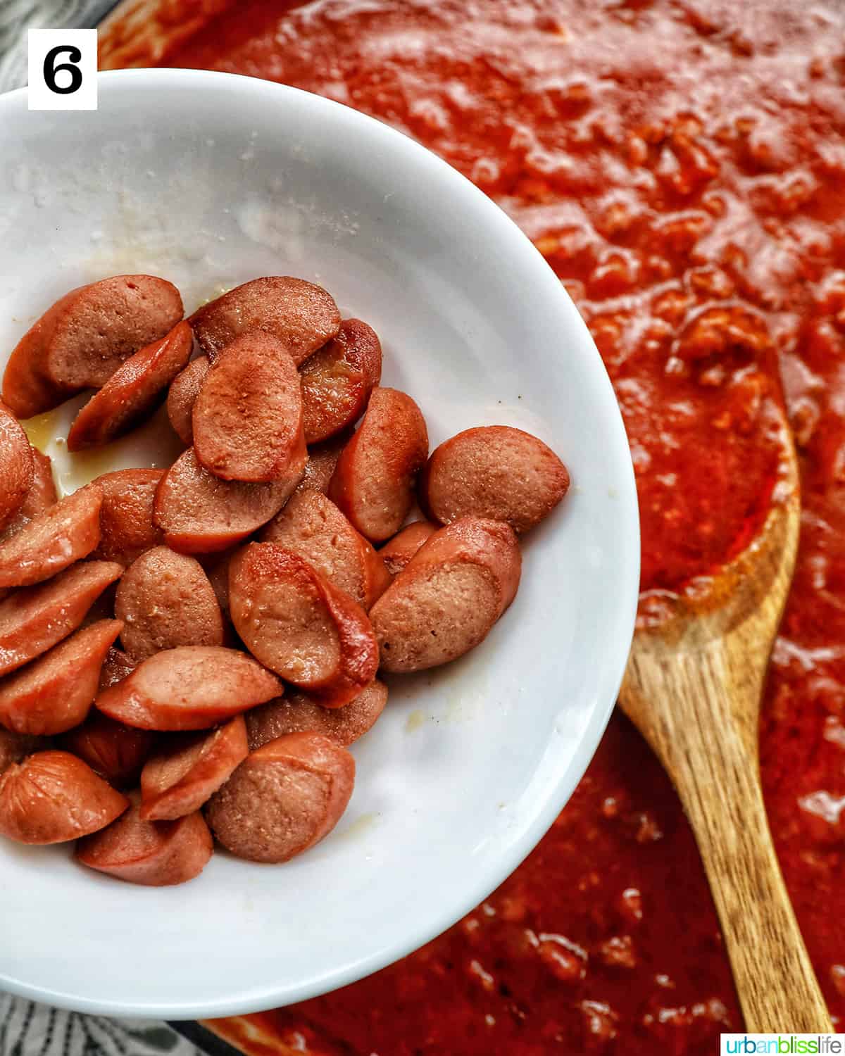 cooked hot dog slices in a white bowl over Filipino spaghetti sauce and a wooden spoon.