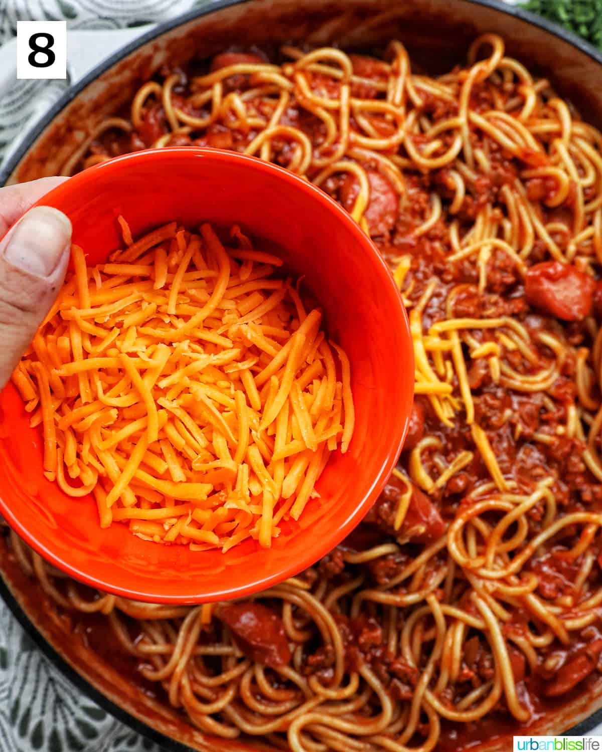 hand holding an orange bowl with shredded cheddar cheese over Filipino spaghetti.