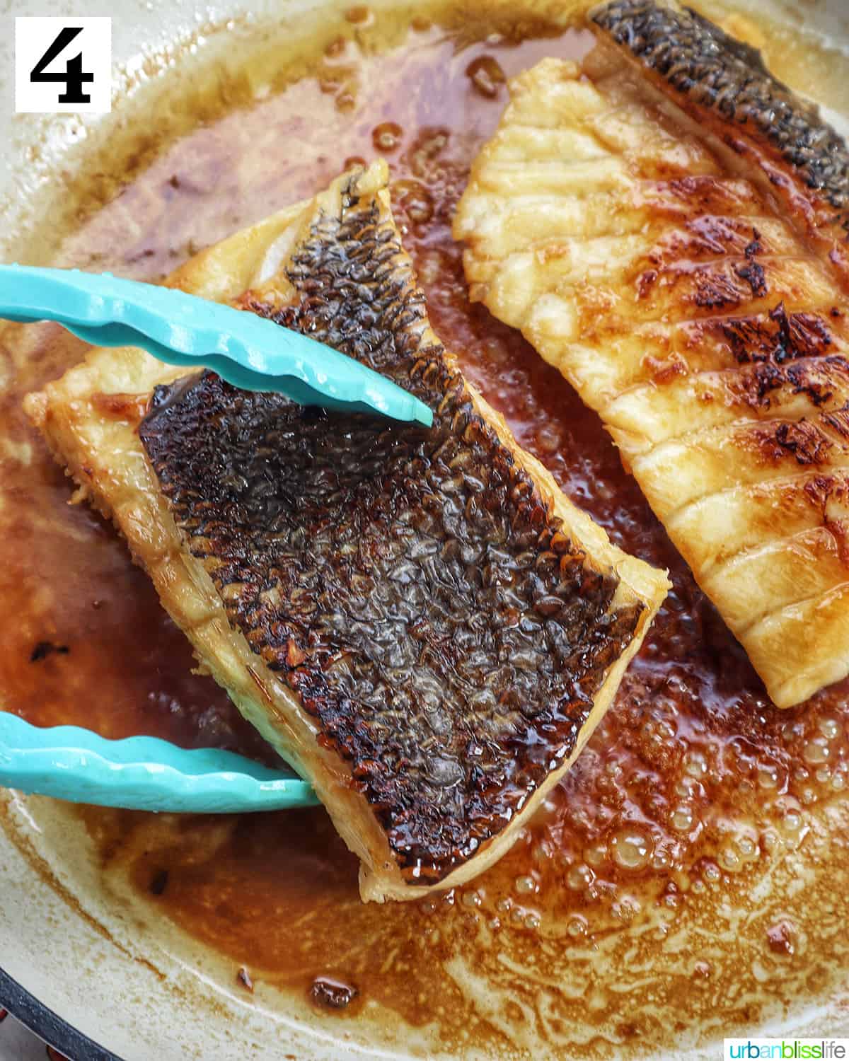 blue tongs flipping over a sea bass fillet next to another fillet in a pan cooking.