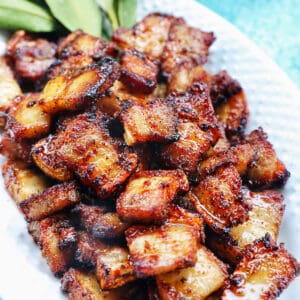 pile of air fryer pork belly bites on a white serving platter with bay leaves on a blue background.