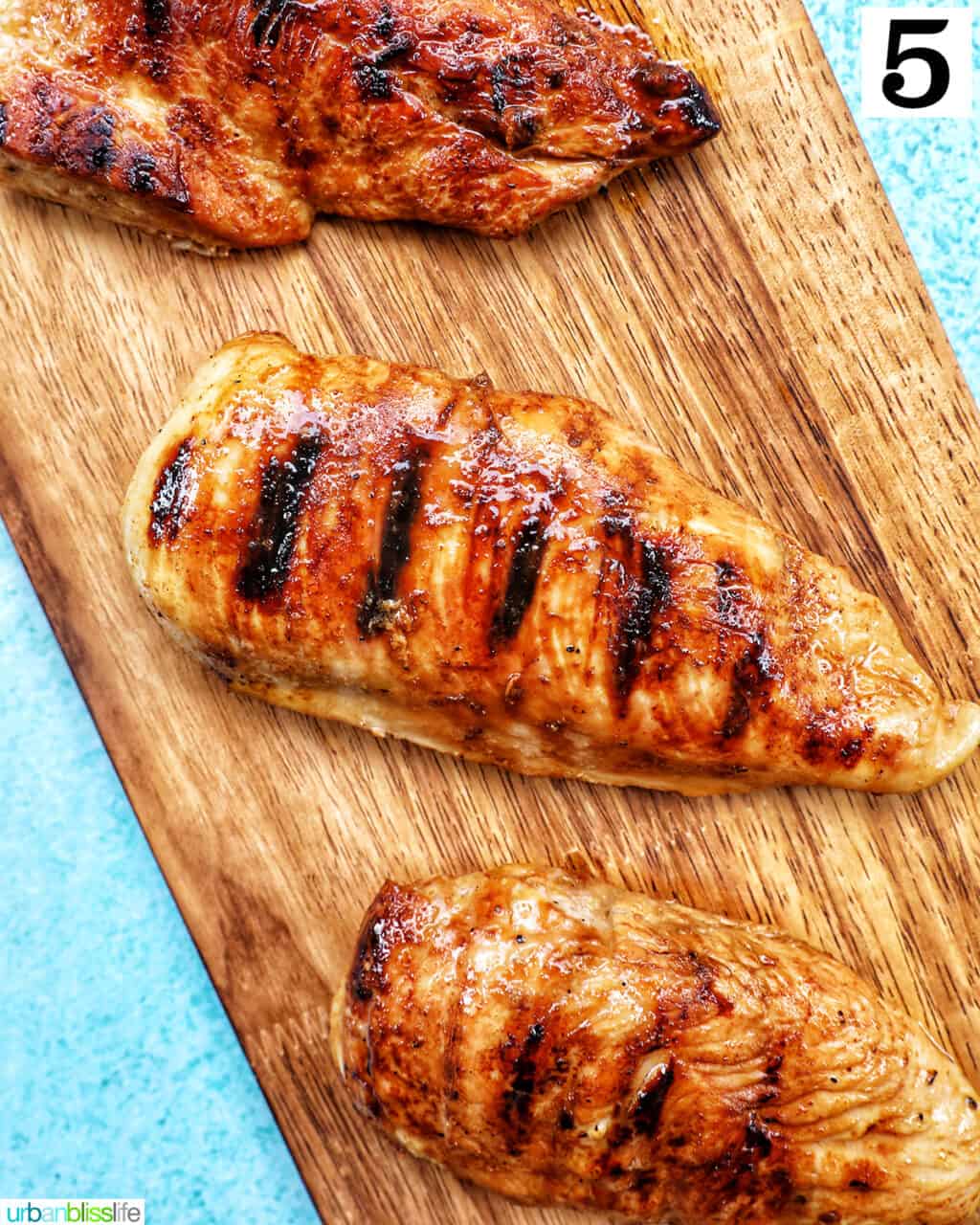 three grilled teriyaki chicken breasts on a wooden board with blue background.