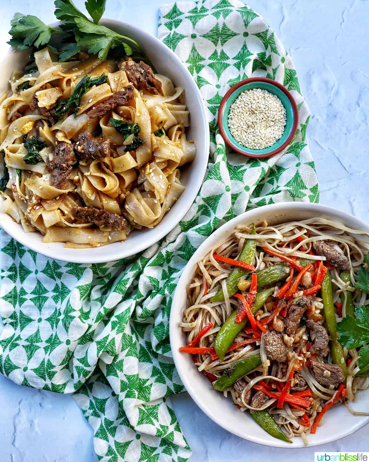 pad see ew in a white bowl above pad thai in a white bowl, both on a green patterned napkin with side of sesame seeds.