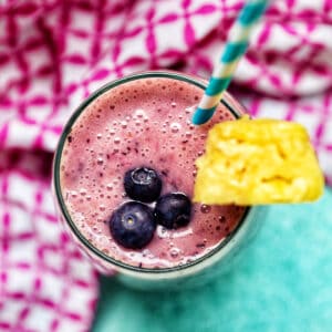 glass of Blueberry Pineapple Smoothie with pineapple wedge garnish, blue and white stripe straw, and pink and white napkin.