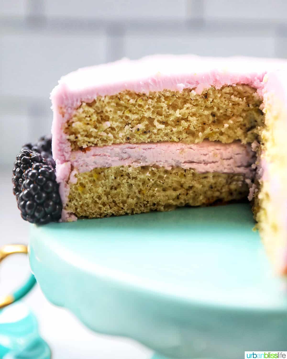 slice of earl grey lavender cake on a purple plate with blackberries with cake on an aqua cake pedestal in the background.