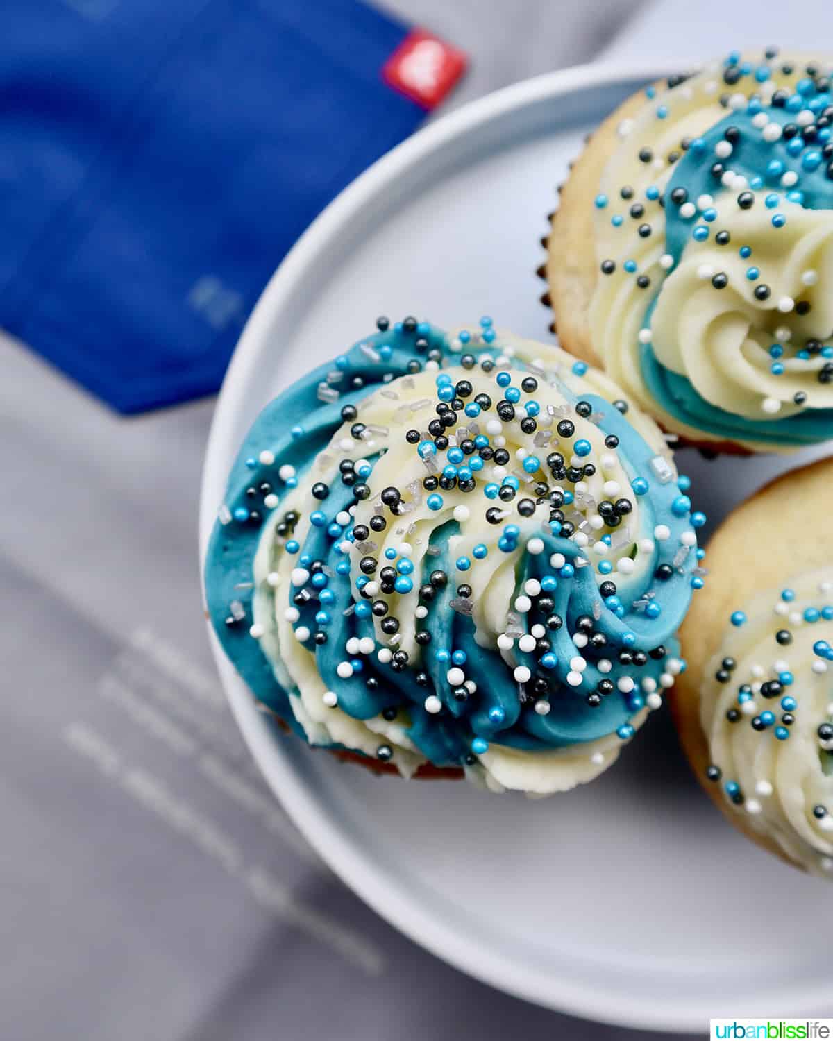 swirls of white and blue vanilla buttercream frosting with white and blue sprinkles on vanilla cupcakes