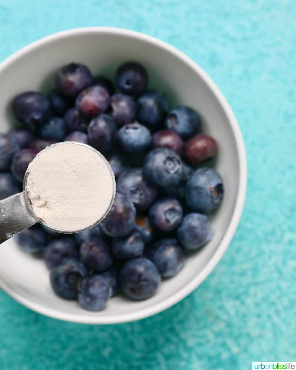 spoon full of flour over a white bowl full of blueberries.
