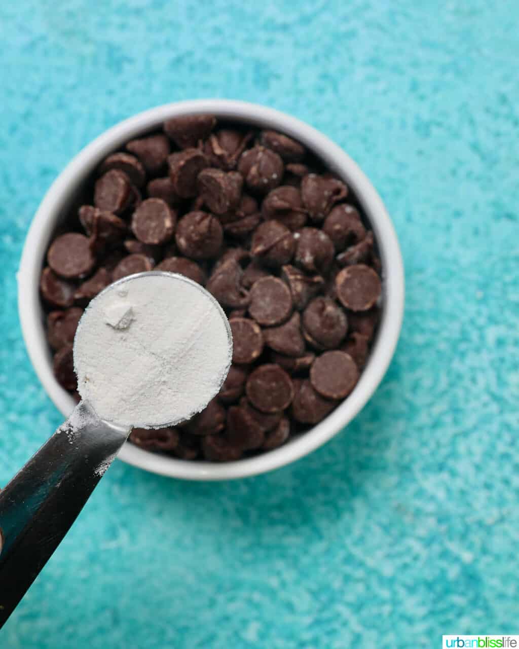 spoonful of flour over a bowl full of chocolate chips on a blue background.