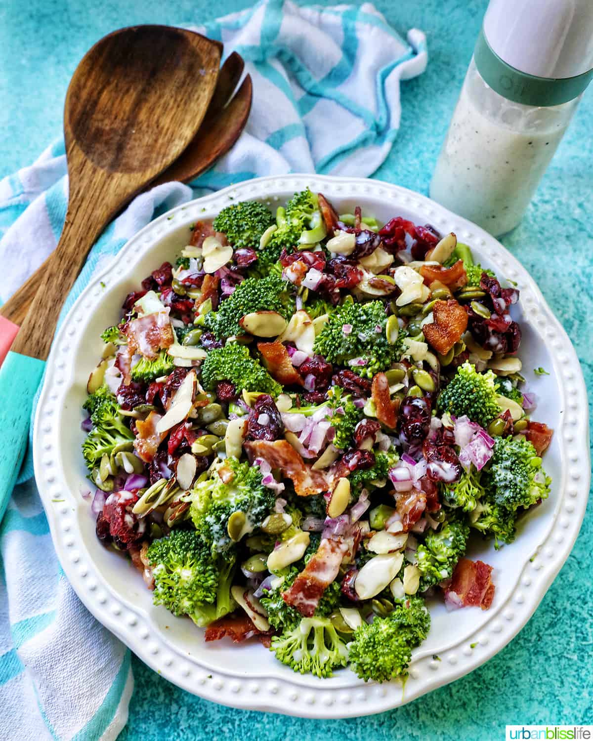 white bowl with broccoli crunch salad with dressing on the side and set of wooden serving utensils on a blue background.