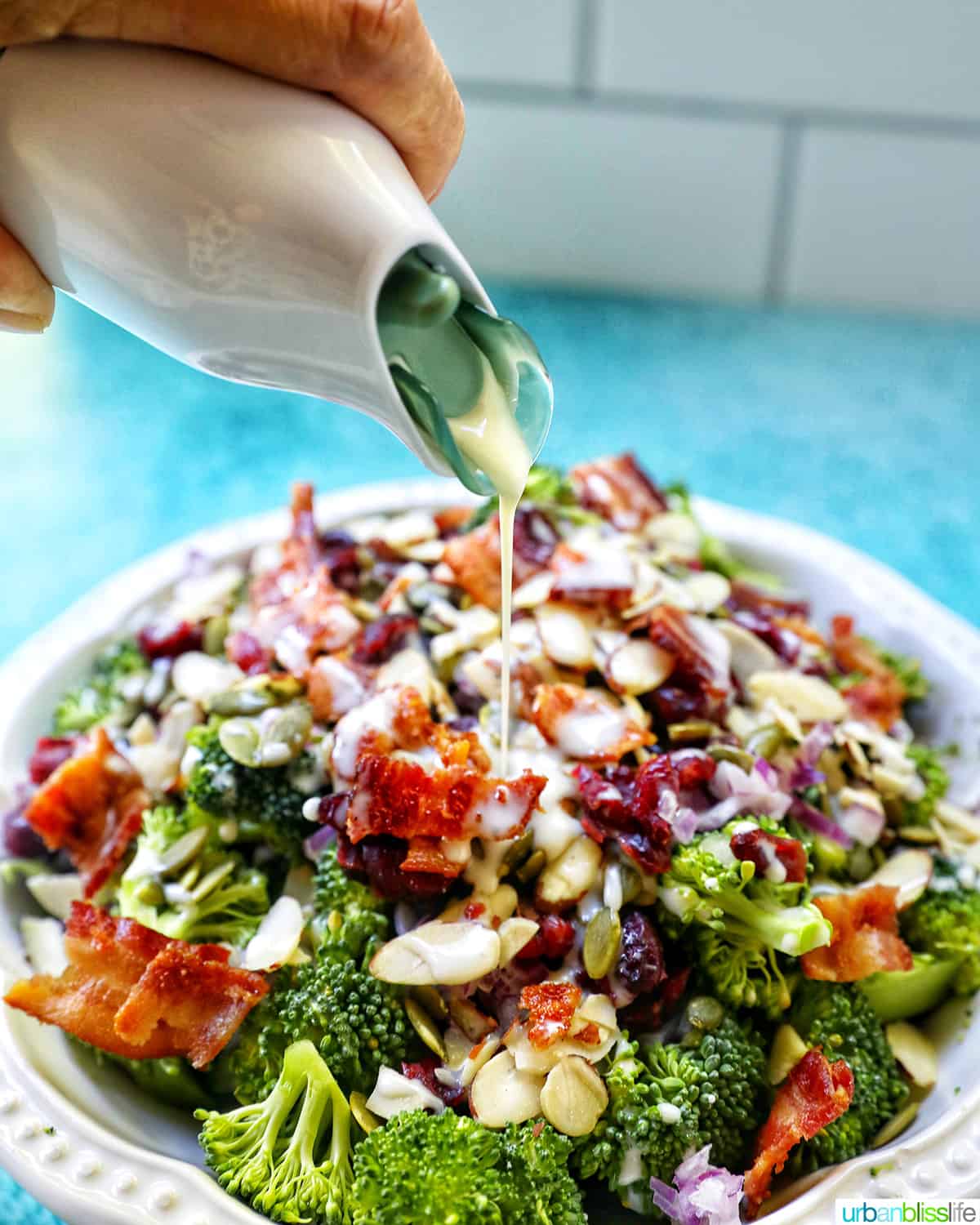 Hand pouring dressing over a white bowl with broccoli crunch salad.