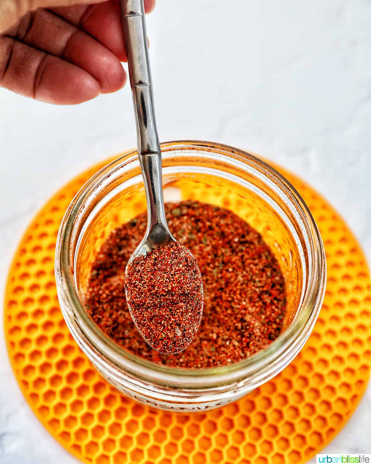 hand holding a spoon in a jar of homemade taco seasoning on a yellow mat.