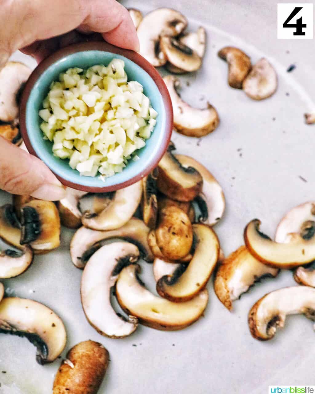 hand holding a small bowl of minced garlic over chopped mushrooms cooking in a pan.