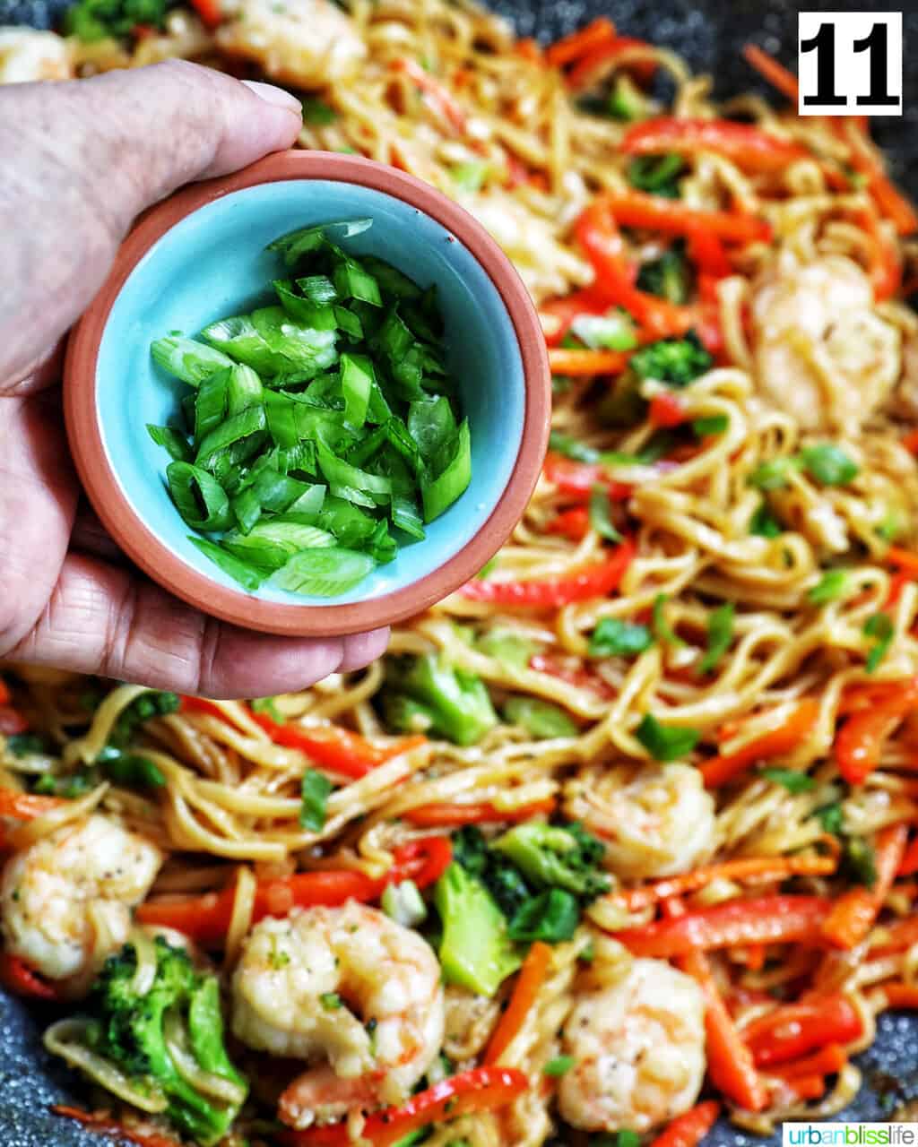hand holding a bowl of chopped green onions over a wok of bowl of prawn chow mein noodles.