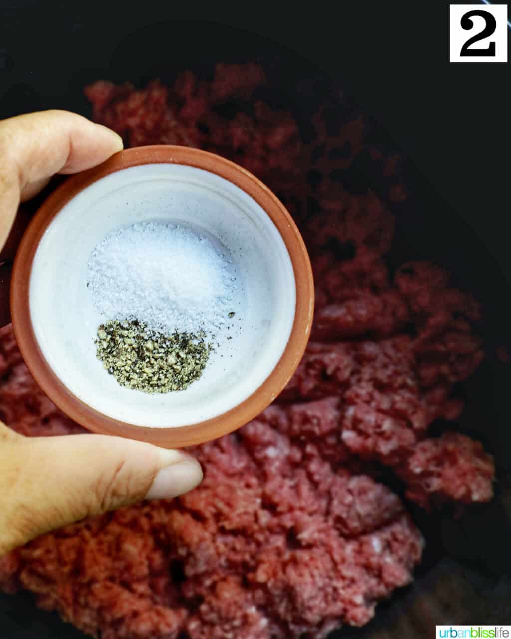 hand holding a small bowl of salt and pepper over ground beef in an air fryer basket.