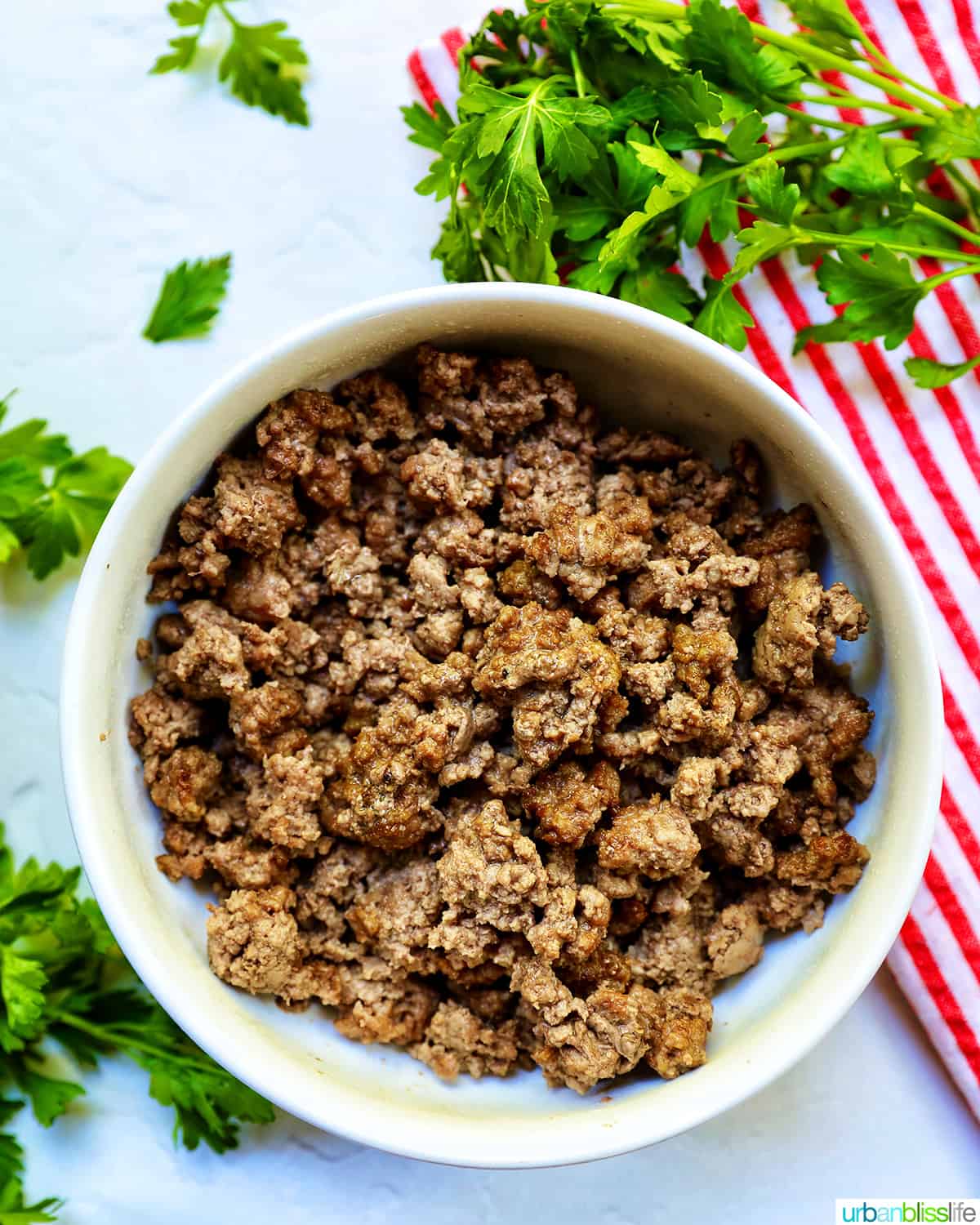 cooked ground beef in a white bowl with parsley sprigs around it and a red and white striped towel.