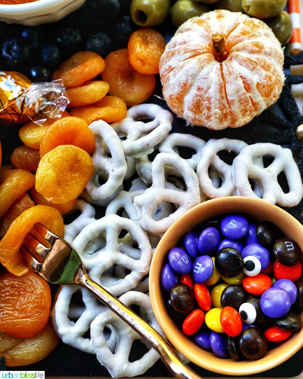 close up of white chocolate covered pretzels, M & Ms, dried apricots and oranges for a Halloween charcuterie board.