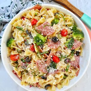 Italian bowtie pasta salad with salami, olives, tomatoes, broccoli, and parmesan cheese in a white bowl with title text that reads "Italian Bow Tie Pasta Salad."