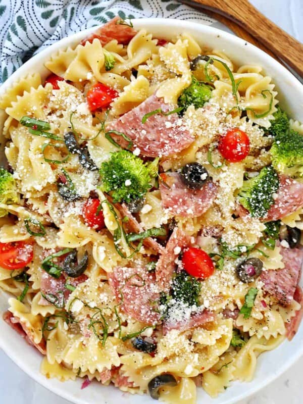 Italian bowtie pasta salad with salami, olives, tomatoes, broccoli, and parmesan cheese in a white bowl with title text that reads "Italian Bow Tie Pasta Salad."