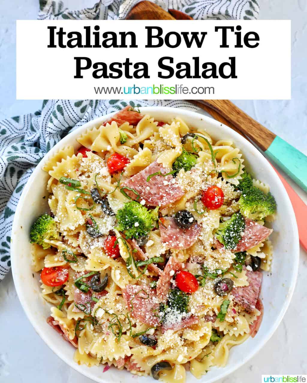 Italian bowtie pasta salad with salami, olives, tomatoes, broccoli, and parmesan cheese in a white bowl with title text that reads "Italian Bow Tie Pasta Salad."