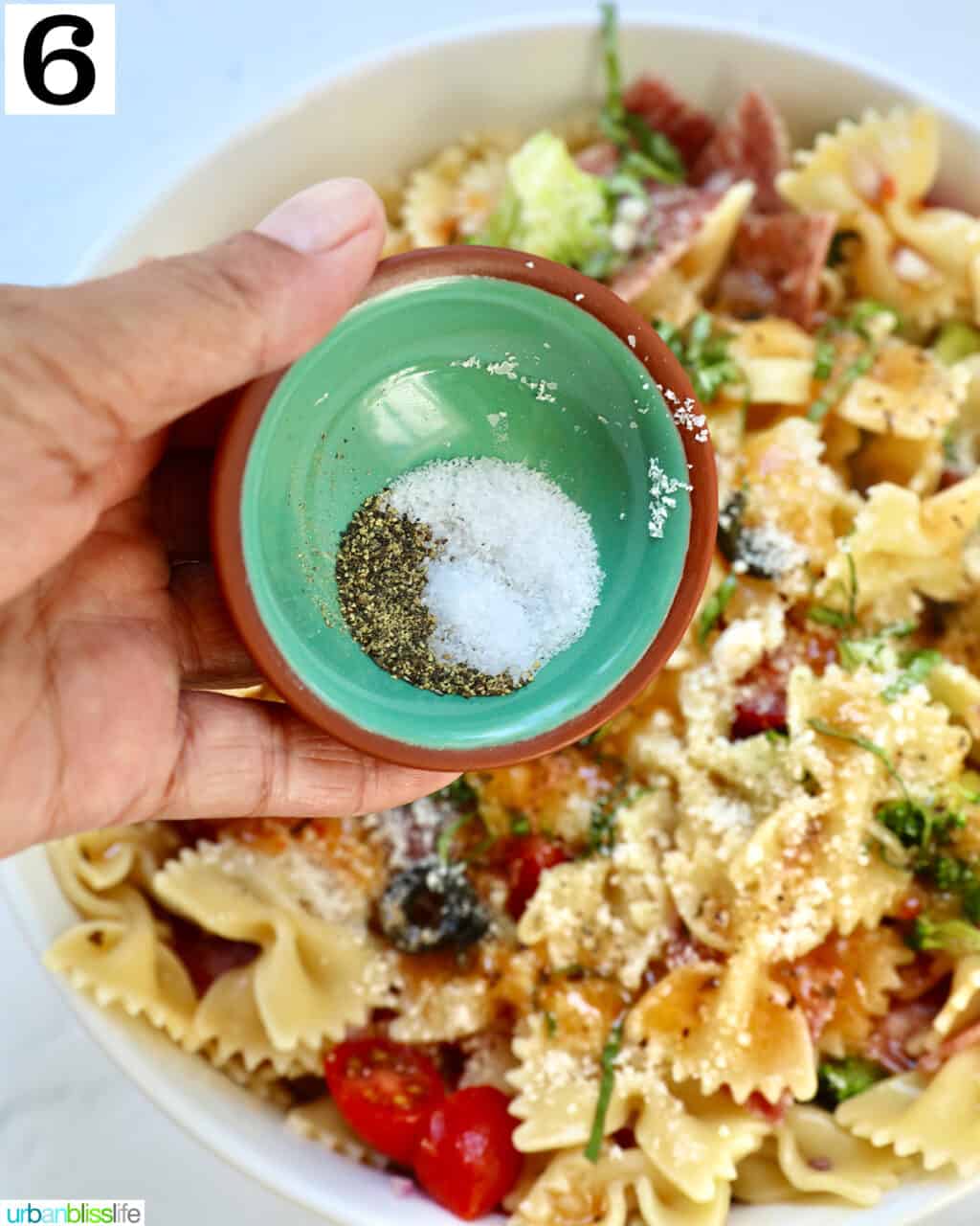 hand holding a small bowl of salt and pepper over a large bowl of Italian pasta salad.