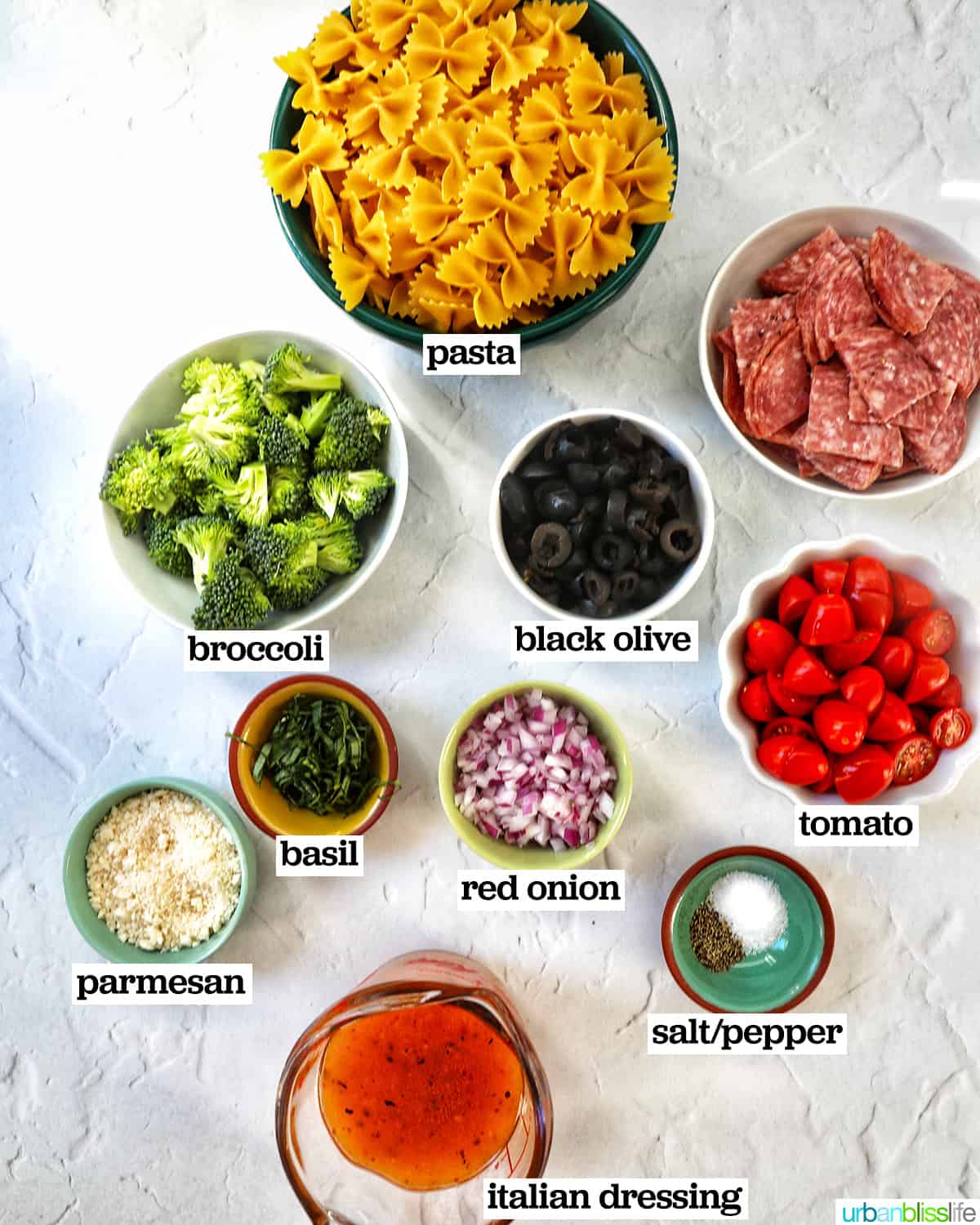 Bowls of ingredients to make italian bowtie pasta salad: tomatoes, red onions, broccoli, pasta, salami, and black olives.
