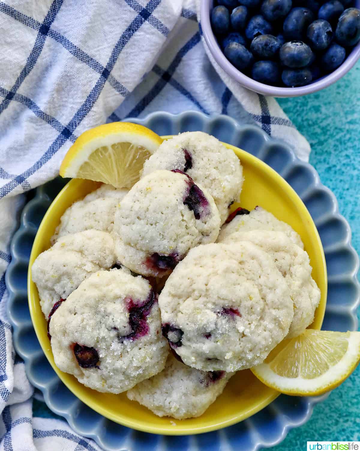 lemon blueberry cookies on a yellow and blue plate with lemon slices, bowl of blueberries, and white and blue napkin.