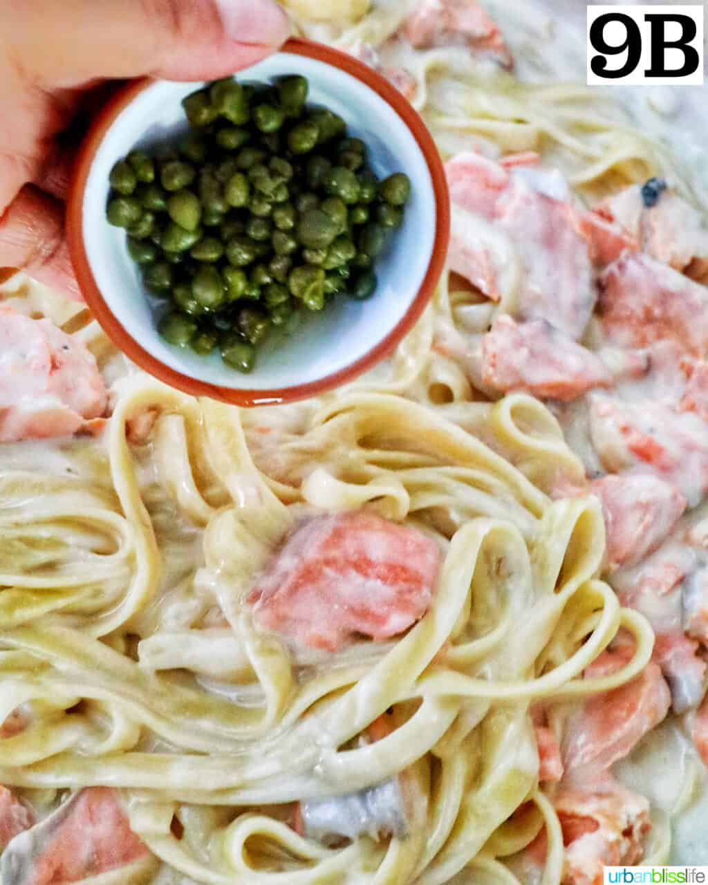 hand holding capers over salmon chunks added to pasta and cream sauce in a skillet.