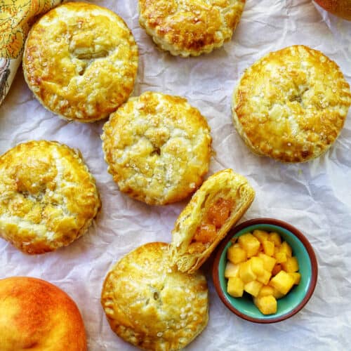several mini peach mango hand pies on crinkled parchment paper with a bowl of diced peaches and mangoes.