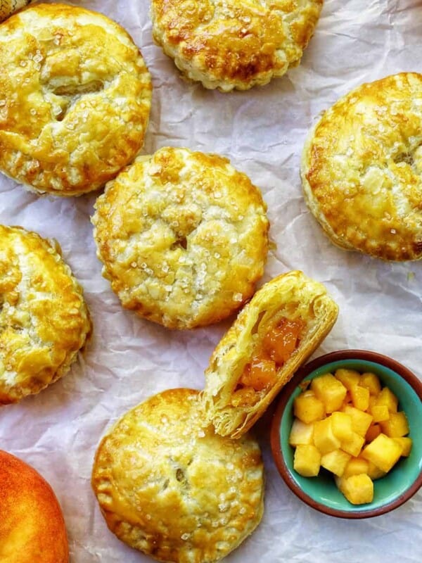 several mini peach mango hand pies on crinkled parchment paper with a bowl of diced peaches and mangoes.