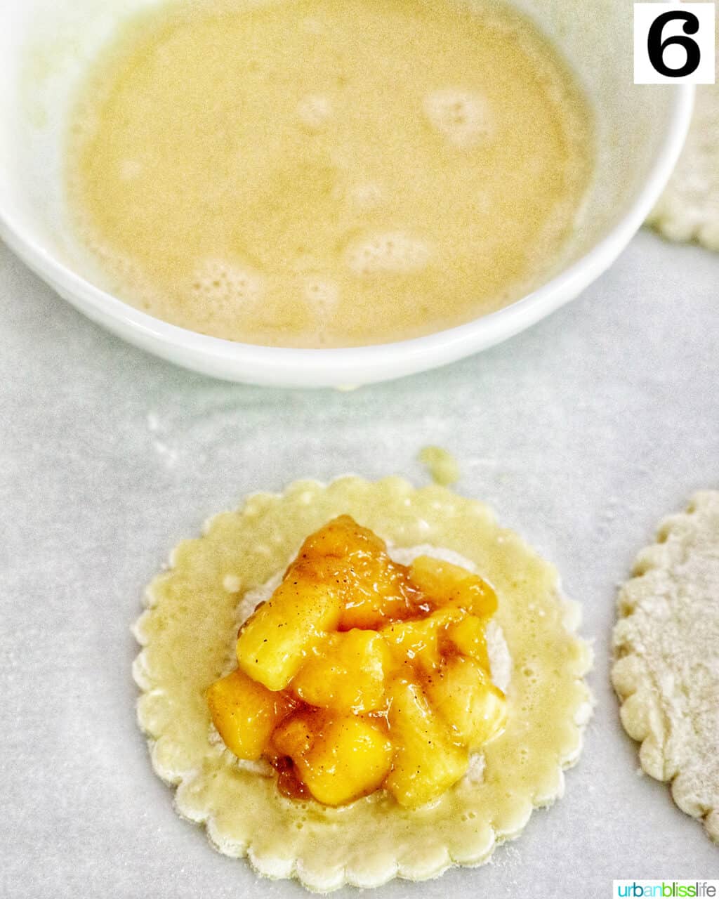 egg wash in a bowl above a puff pastry round with diced peaches and mangoes on top.