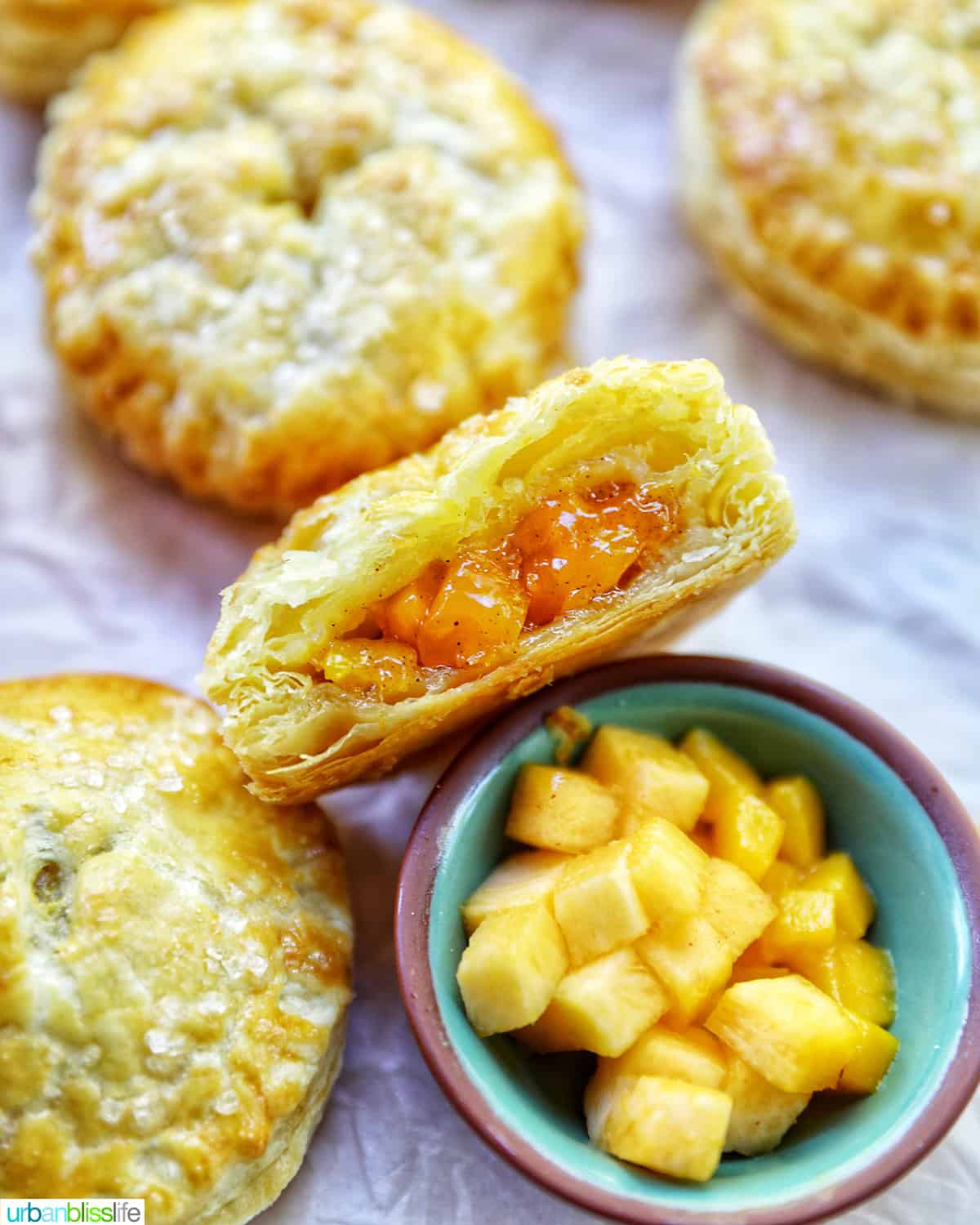 several mini peach mango hand pies on crinkled parchment paper with a bowl of diced peaches and mangoes.