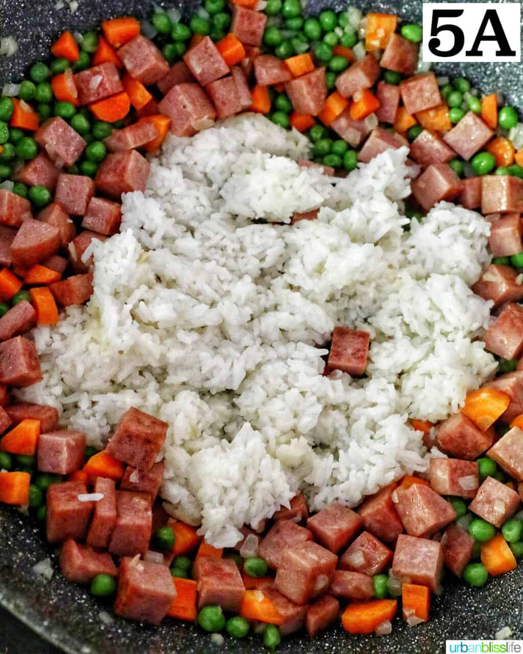 adding rice to cooking cubed spam, garlic, onions, peas, and carrots in a wok.
