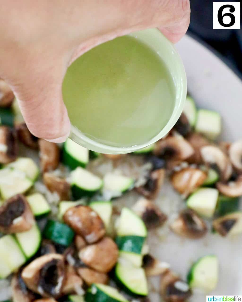 hand holding a bowl of white wine over a saucepan of mushrooms and zucchini.