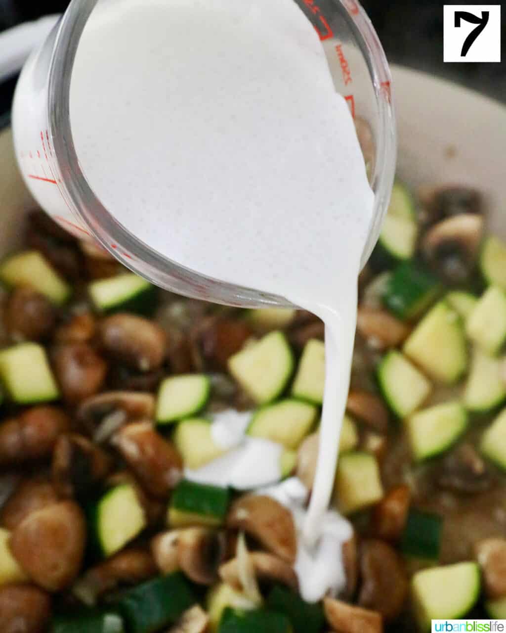 cream pouring out of a measuring cup into a pan of mushrooms and zucchini.