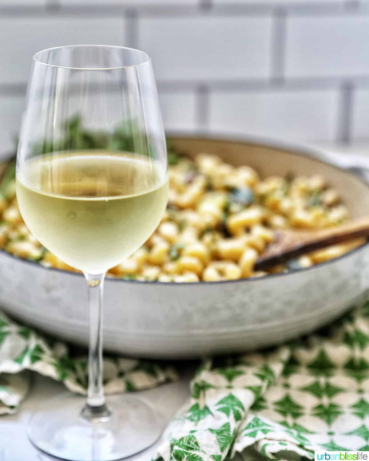 glass of white wine in front of a pan of pasta with a green and white napkin.