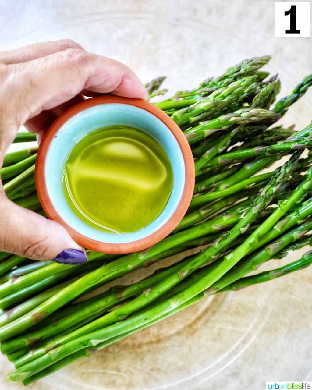 hand holding a bowl of olive oil over asparagus.