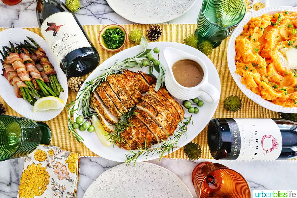 Thanksgiving table with sliced turkey tenderloin and gravy, whipped sweet potatoes, bacon wrapped asparagus, two bottles of wine.