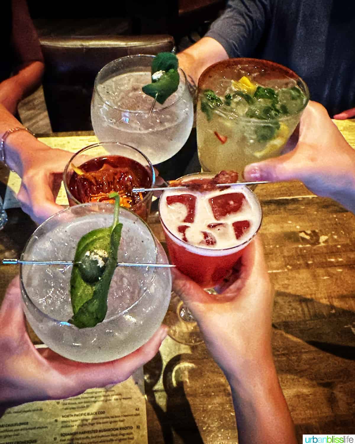five hands holding cocktails up to cheers at the Raven Room in Whistler BC Canada.