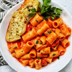 white bowl with rigatoni with vodka sauce and side of garlic bread and parsley.