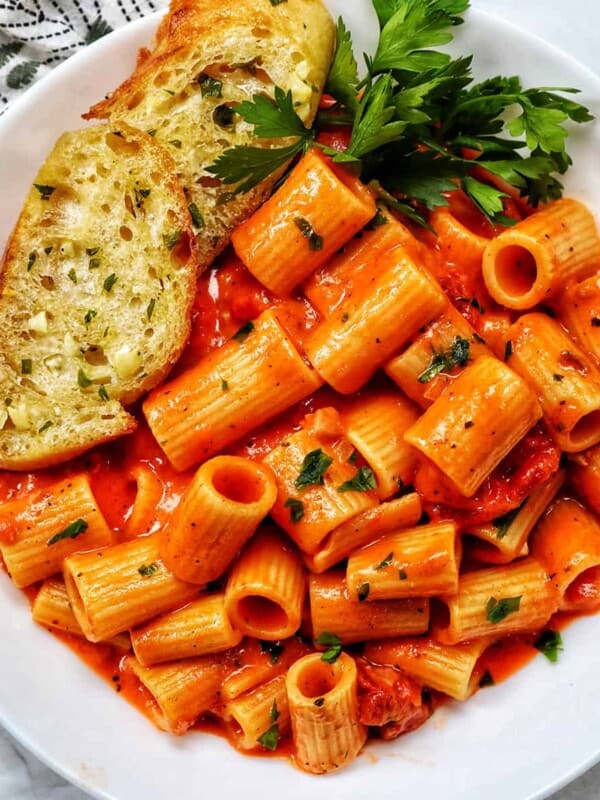white bowl with rigatoni with vodka sauce and side of garlic bread and parsley.