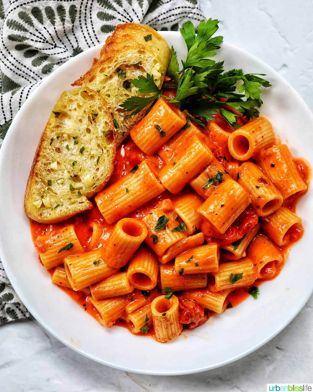 white bowl with rigatoni with vodka sauce and side of garlic bread and parsley.