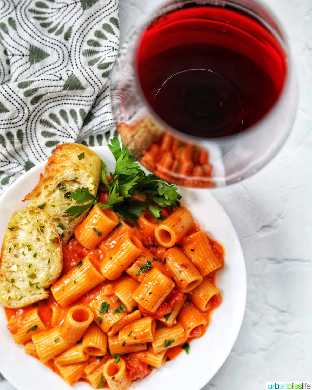 glass of red wine and white bowl with rigatoni with vodka sauce and side of garlic bread and parsley.