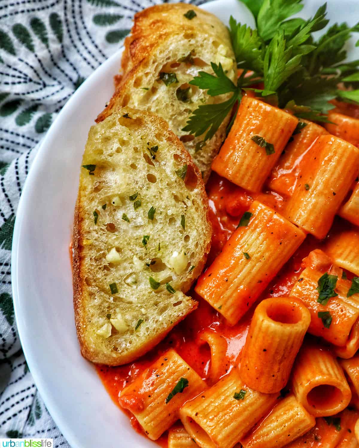 white bowl with rigatoni with vodka sauce and side of garlic bread and parsley.