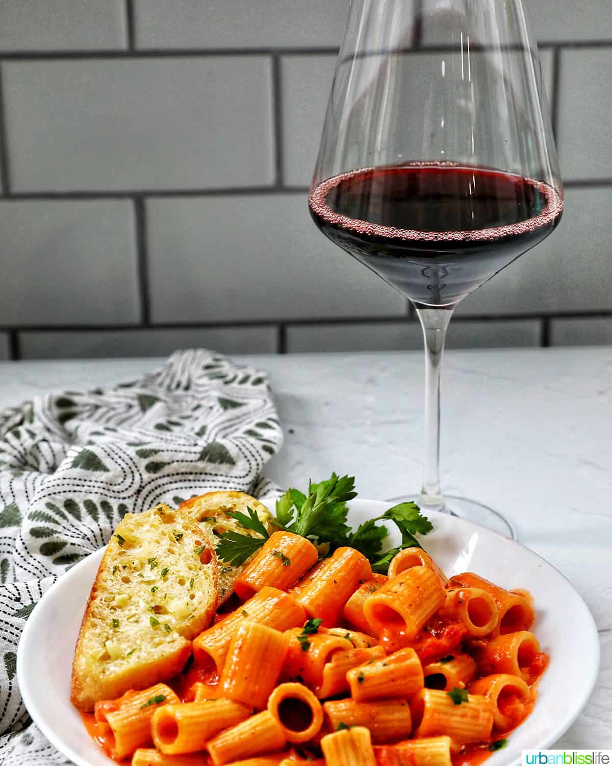 glass of red wine with white bowl with rigatoni with vodka sauce and side of garlic bread and parsley.