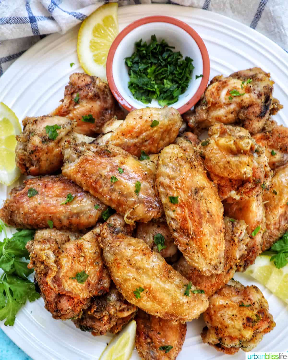 white plate full of salt and pepper wings with side of chopped fresh parsley and lemon wedges.