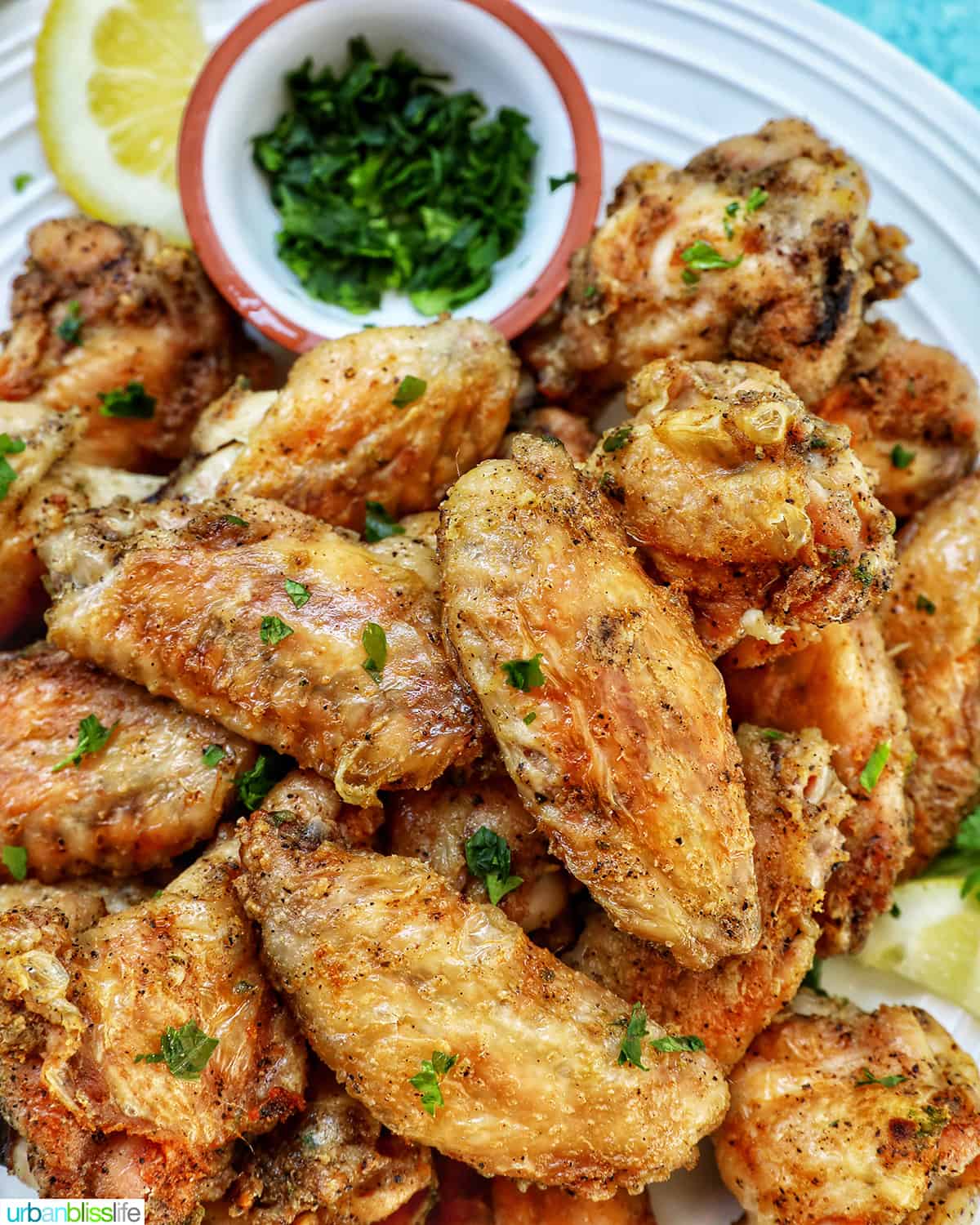 white plate full of salt and pepper wings with side of chopped fresh parsley and lemon wedges.